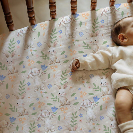 Baby lying on a crib mattress with a bunny pattern, wearing a white outfit.