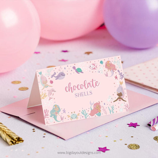 Card with mermaid design and 'chocolate shells' text on a pink envelope with confetti and balloons in the background.